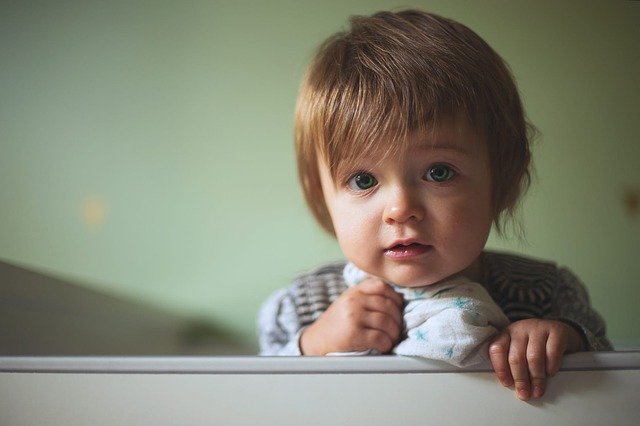 Dieťa s hnedými vlasmi pozerá cez zábradlie z postieľky.jpg